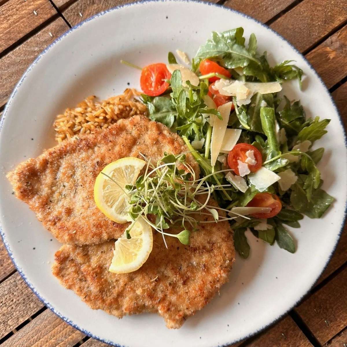 Crispy breaded chicken cutlets garnished with lemon slices and microgreens, served with a fresh arugula salad topped with cherry tomatoes and shaved Parmesan, alongside a portion of seasoned rice pilaf on a rustic ceramic plate.