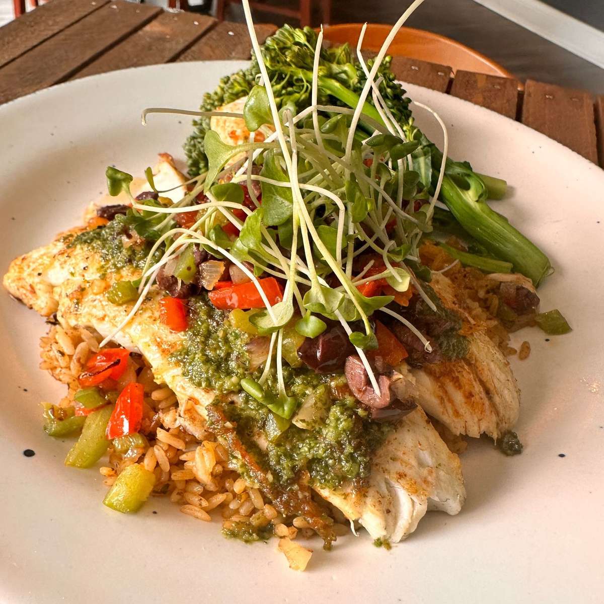 Grilled white fish fillet served over seasoned rice with roasted peppers and olives, topped with chimichurri sauce and fresh microgreens, accompanied by steamed broccolini on a rustic white plate.