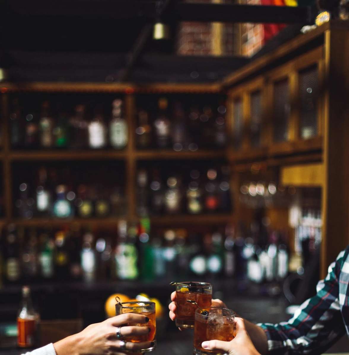 Guests toasting with cocktails at a dimly lit bar, featuring shelves lined with liquor bottles in the background, creating a lively and inviting nightlife atmosphere.
