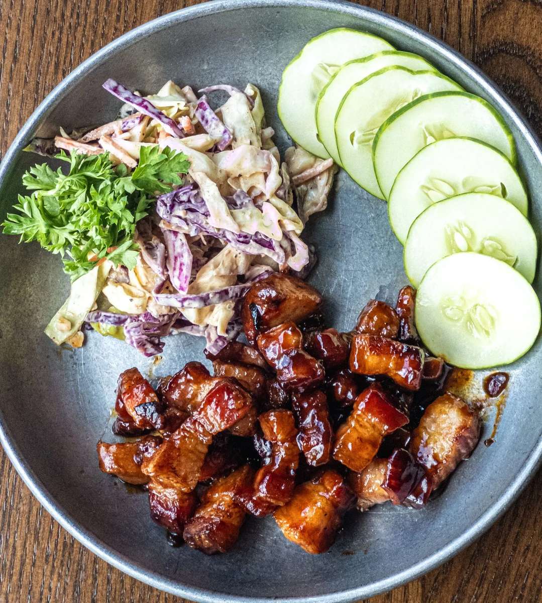 Glazed pork belly bites with a caramelized finish, served alongside creamy coleslaw with purple and green cabbage, fresh cucumber slices, and a parsley garnish on a rustic metal plate.
