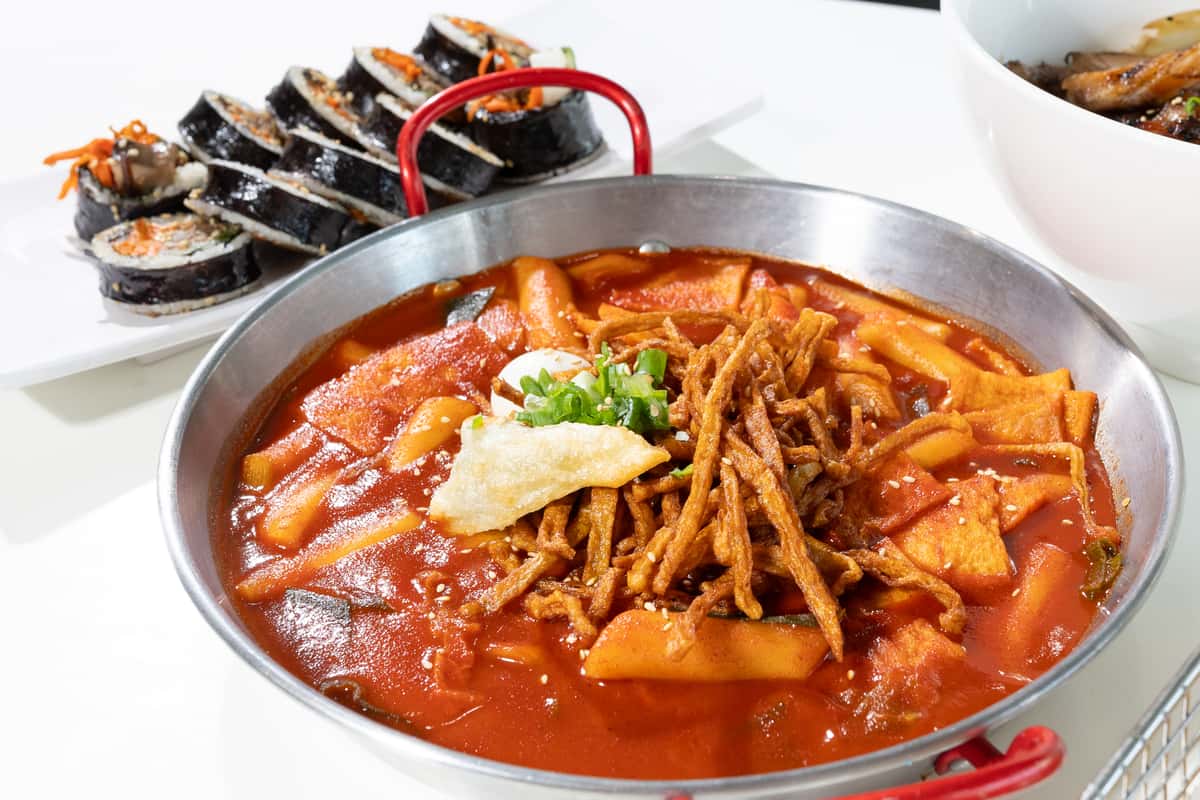 A bowl of spicy Korean tteokbokki topped with fried toppings and green onions, served alongside sliced gimbap rolls.