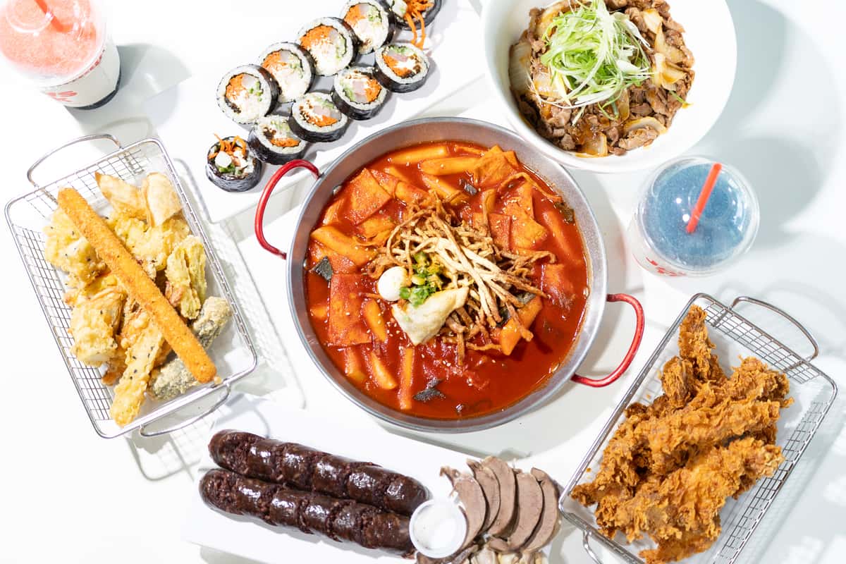 Korean feast with spicy tteokbokki, kimbap rolls, fried chicken, assorted tempura, soondae, bulgogi rice bowl, and colorful drinks.