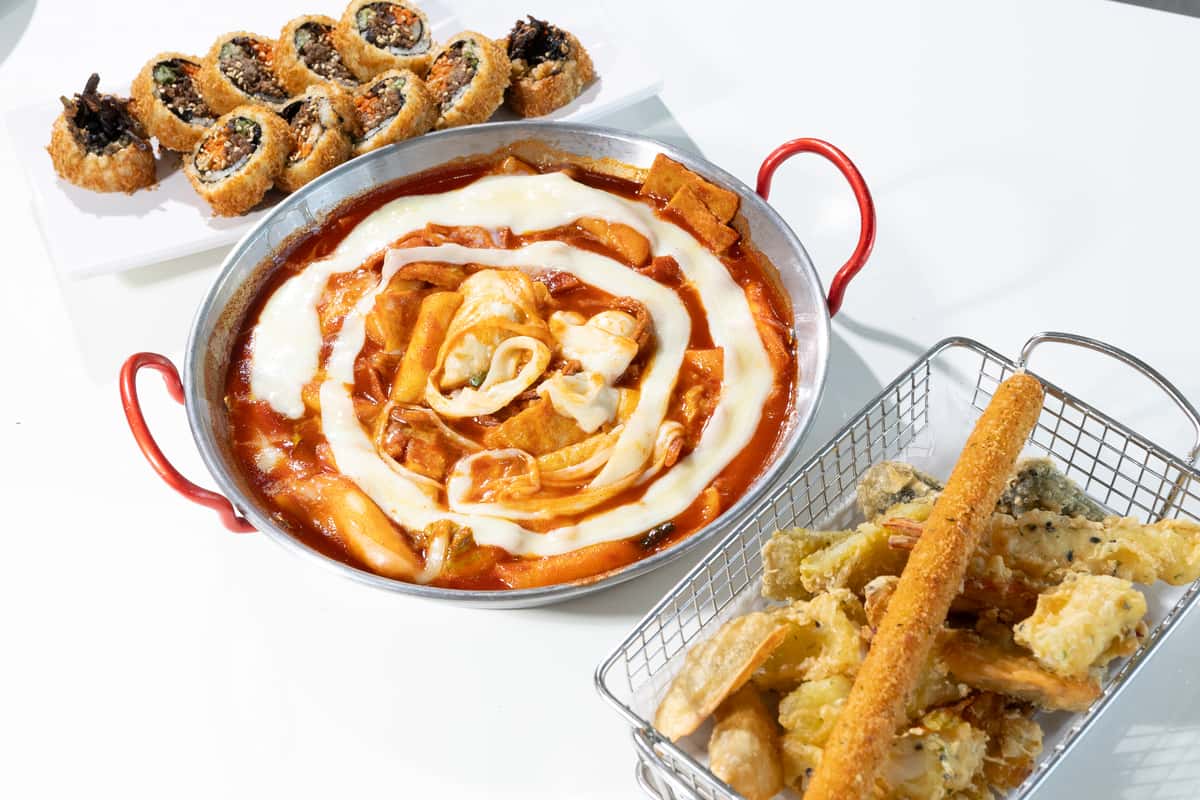Korean tteokbokki topped with melted cheese, served with fried gimbap and assorted tempura on a white table.