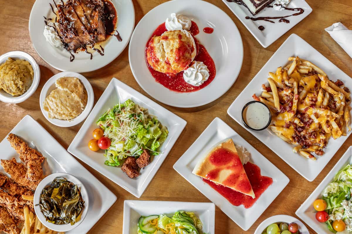Overhead view of a Southern comfort food spread on a wooden table, featuring fried chicken tenders with fries and collard greens, cheesy bacon-covered fries with ranch dip, garden salad with cherry tomatoes and croutons, creamy mashed potatoes with gravy, golden biscuits, and rich entrees including BBQ-glazed ribs and meatloaf. Desserts include a strawberry sauce-topped biscuit with whipped cream, a slice of cheesecake, and a brownie drizzled with chocolate syrup.