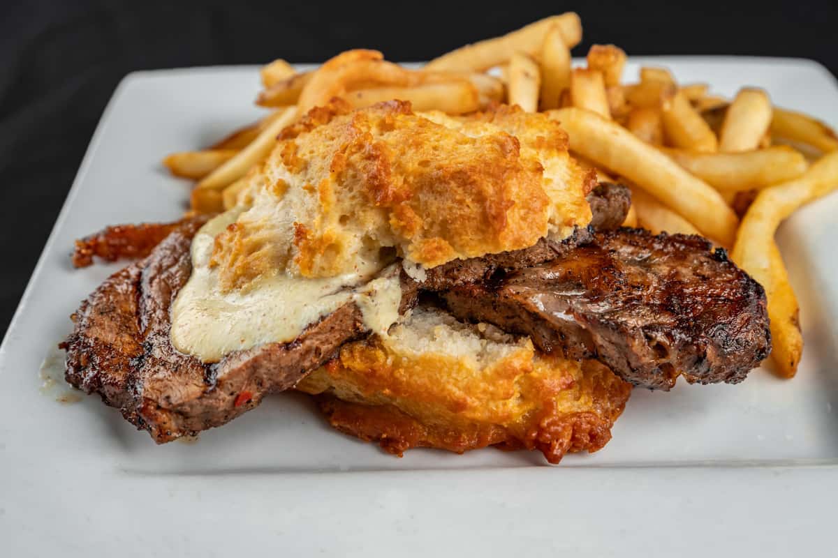Close-up of a hearty Southern-style steak biscuit sandwich served with golden French fries, featuring grilled beef steak layered with melted cheese between a flaky, cheddar-topped biscuit on a white plate.