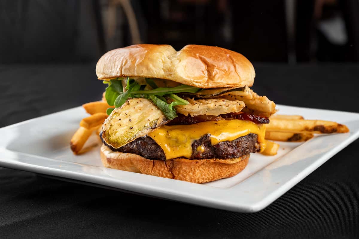 Gourmet cheeseburger topped with crispy kettle-cooked potato chips, fresh greens, and melted cheddar on a toasted brioche bun, served with golden French fries on a square white plate