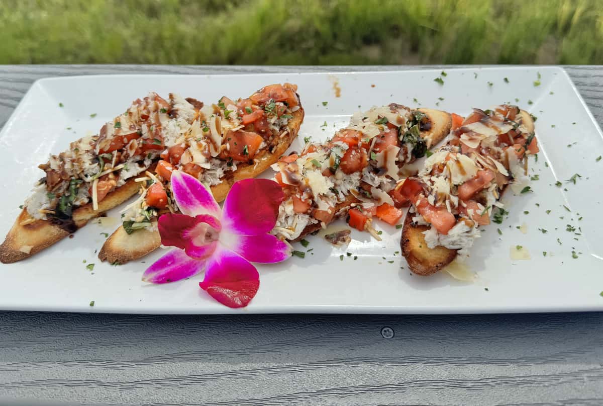 Toasted baguette slices topped with tomato bruschetta, shredded crab, shaved parmesan, and balsamic drizzle, garnished with orchid.