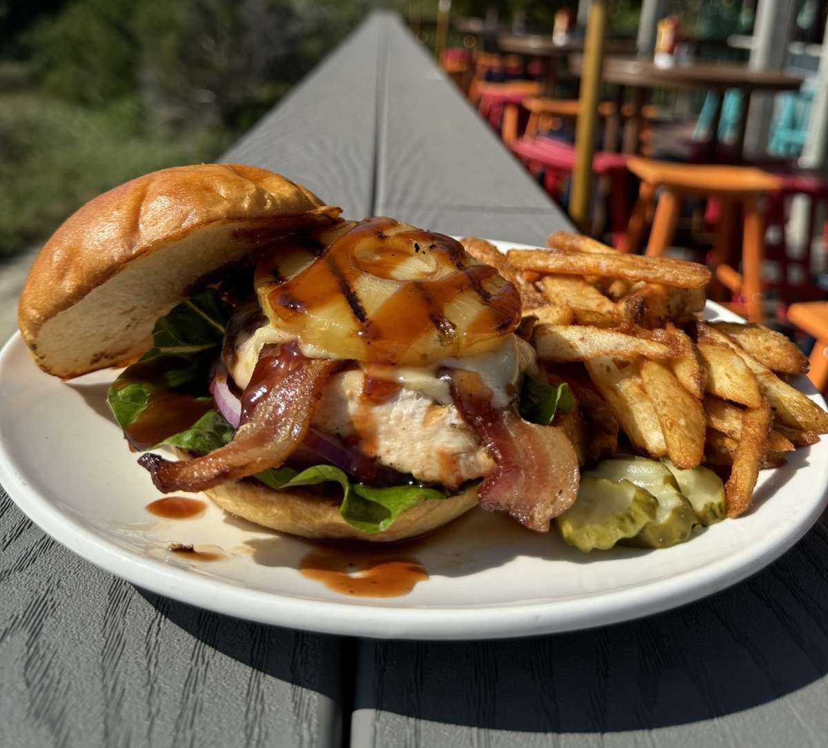 Grilled chicken sandwich with bacon, pineapple, lettuce, and sauce served with fries and pickles on a sunny outdoor patio.