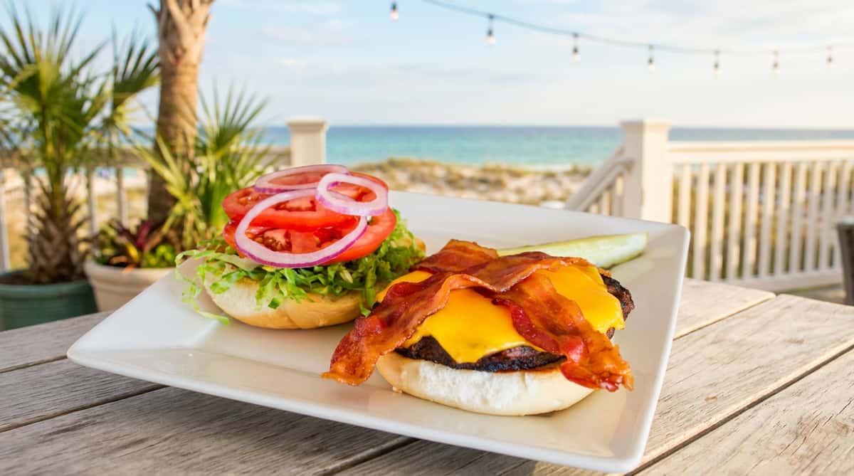 Bacon cheeseburger with shredded lettuce, tomato, red onion, and a pickle spear on a white plate.