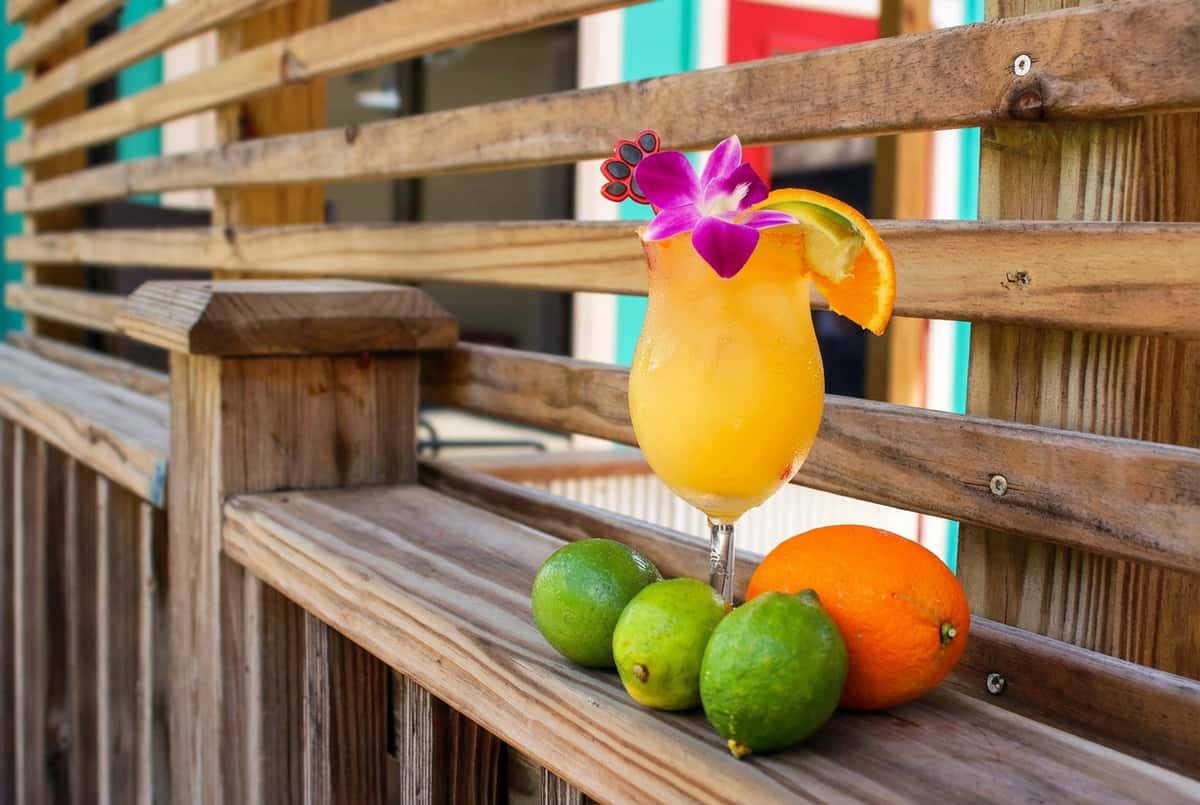Yellow cocktail with purple orchid, orange slice, and 3 limes on a wooden slat railing.