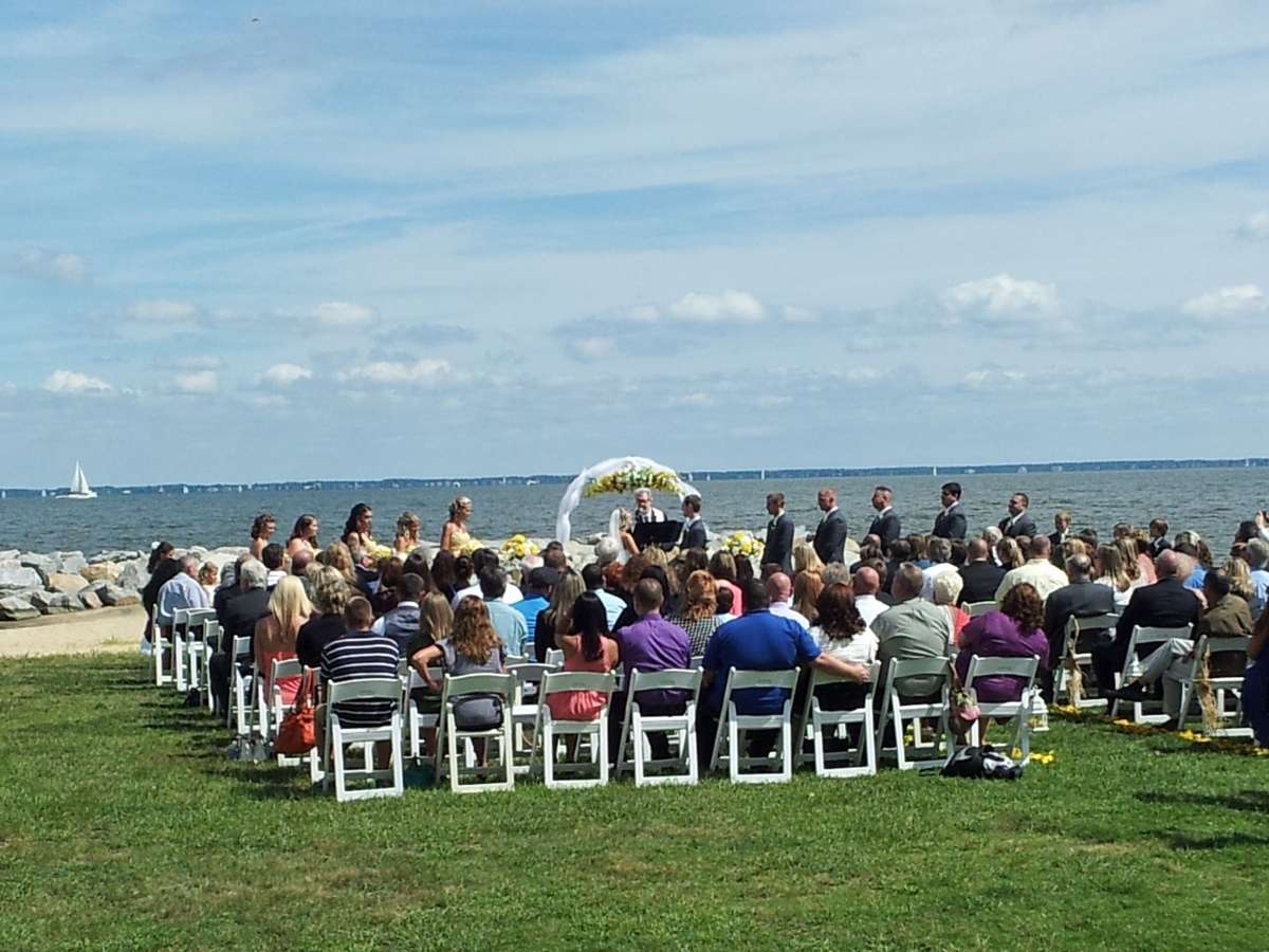 Wedding ceremony with a large group of people taking place outdoors on the grass by the water