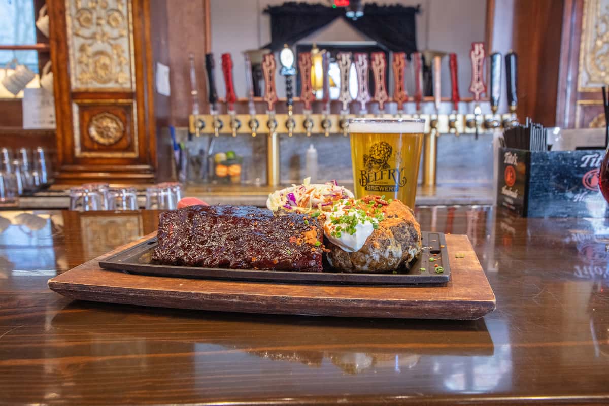 BBQ ribs glazed with sauce served on a wooden board with coleslaw and sides, set on a brewery bar with beer taps in the background