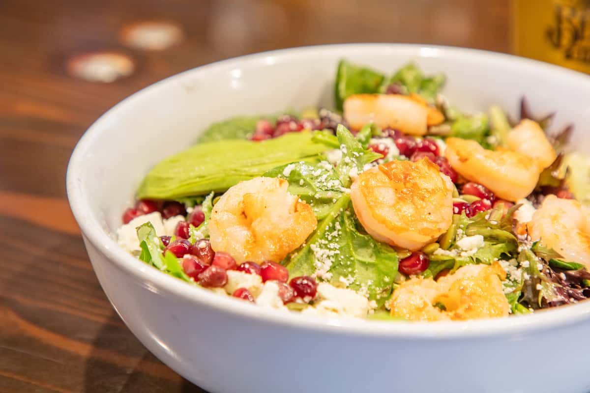 Fresh mixed green salad topped with seared scallops, pomegranate seeds, and grated cheese in a white bowl at a restaurant