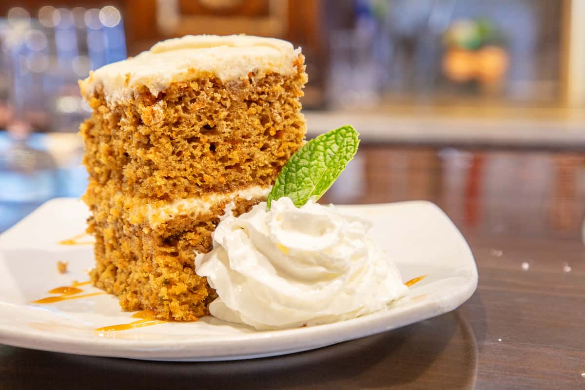Moist carrot cake slice with cream cheese frosting served with whipped cream and caramel drizzle on a dessert plate at a cozy restaurant