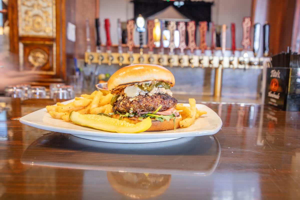 Gourmet burger with blue cheese crumbles, jalapeños, and greens on a toasted bun, served with fries at a brewery bar
