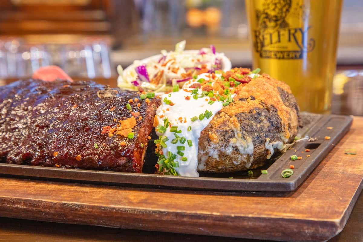 BBQ ribs platter with baked potato and creamy coleslaw served on a wooden board at a craft brewery restaurant with bar taps in the background