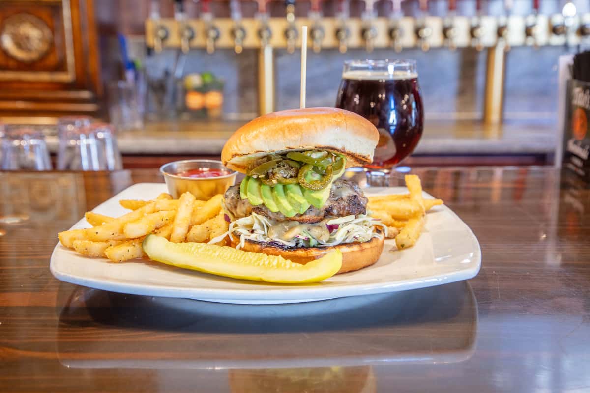 Southwest-style cheeseburger with guacamole, lettuce, and tomato served with fries and a pickle alongside a glass of craft beer on a brewery bar counter