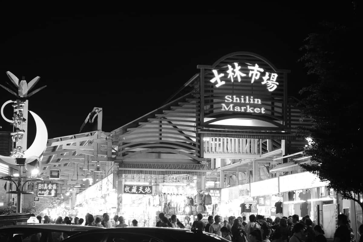 Black-and-white nighttime view of the illuminated Shilin Market entrance and surrounding crowds in Taipei, Taiwan.