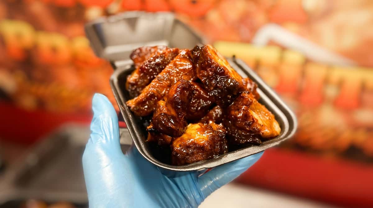 A gloved hand holds a black take-out container filled with saucy, dark glazed chicken wings.