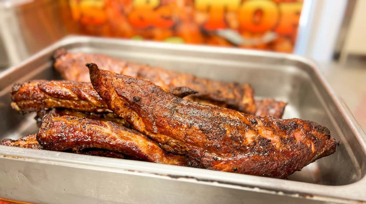 Slow-cooked ribs with dark, crispy char, glistening with sauce in a metal serving pan.