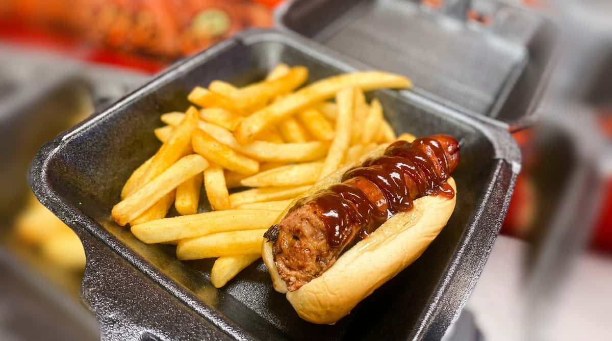 Hot dog with BBQ sauce in a bun, served with golden-brown french fries in a black to-go container.