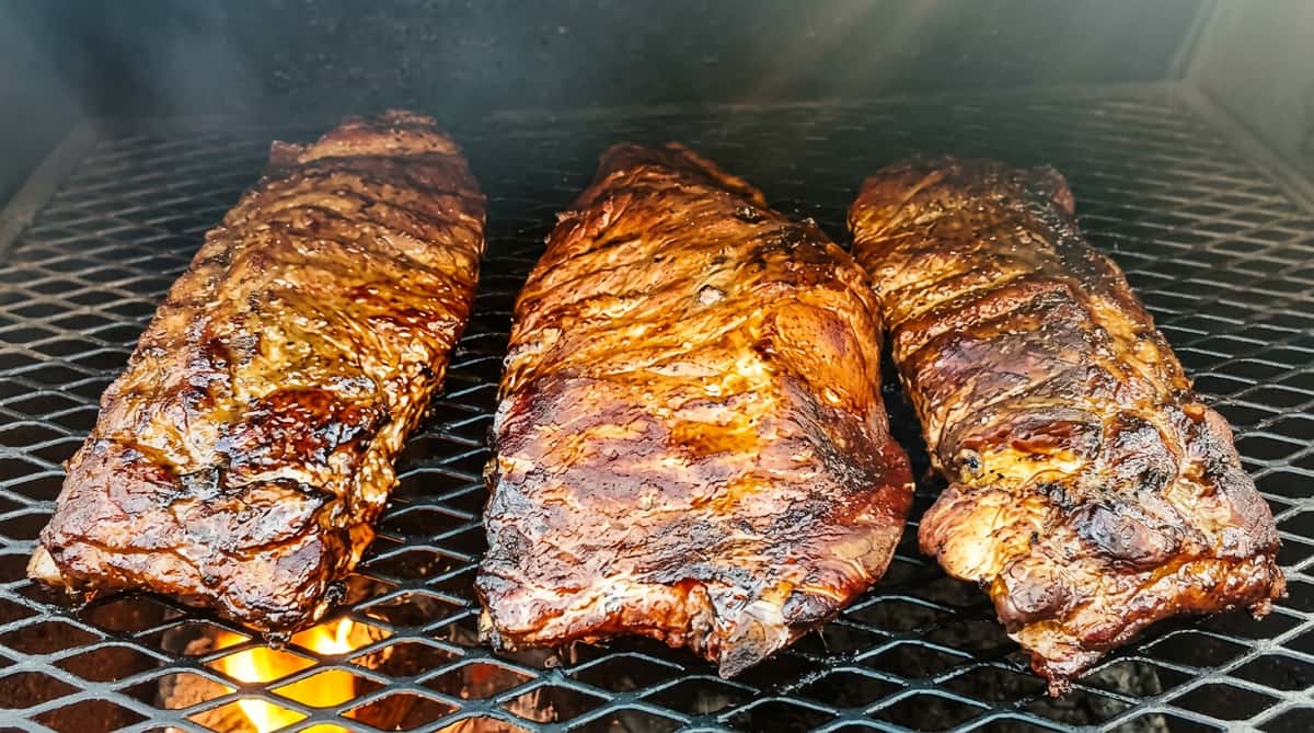 Three large racks of ribs cooking on a grill, with visible flames beneath.