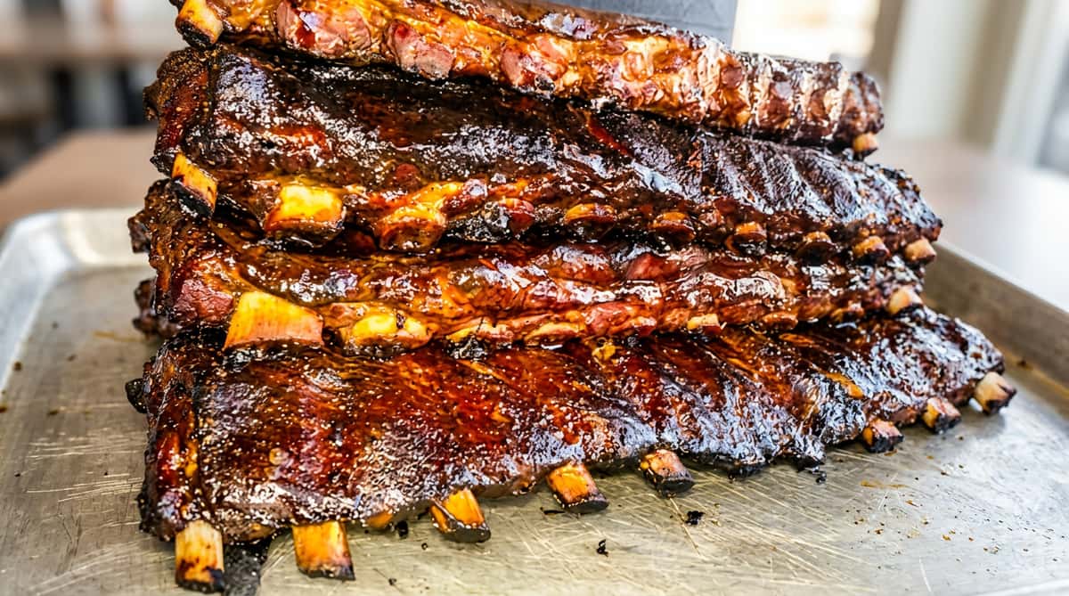 Three racks of smoked BBQ ribs, glistening with a dark, rich sauce, are stacked on a table.