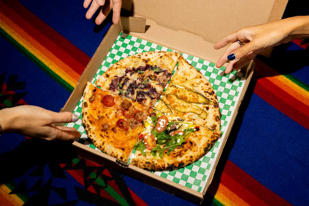 Takeout pizza with assorted toppings—pepperoni and sausage, veggie, arugula and cherry tomatoes, and white pizza slices—served in an open box as hands reach in, set on a colorful patterned tablecloth.