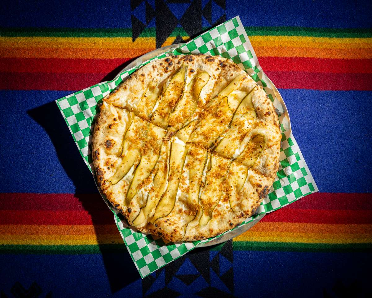 Wood-fired white pizza topped with thinly sliced zucchini, melted cheese, and crispy crust on a colorful textile backdrop