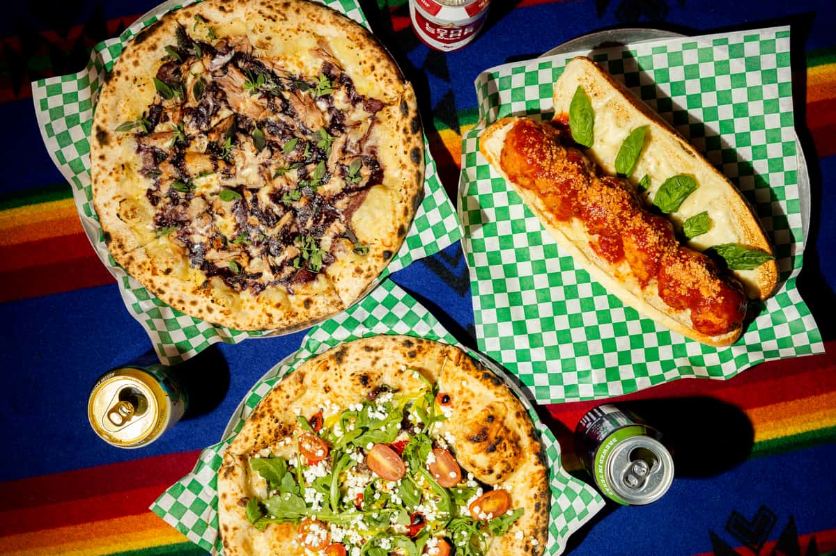 Wood-fired pizzas with gourmet toppings and a meatball sub with marinara, served with canned drinks on a colorful tablecloth.