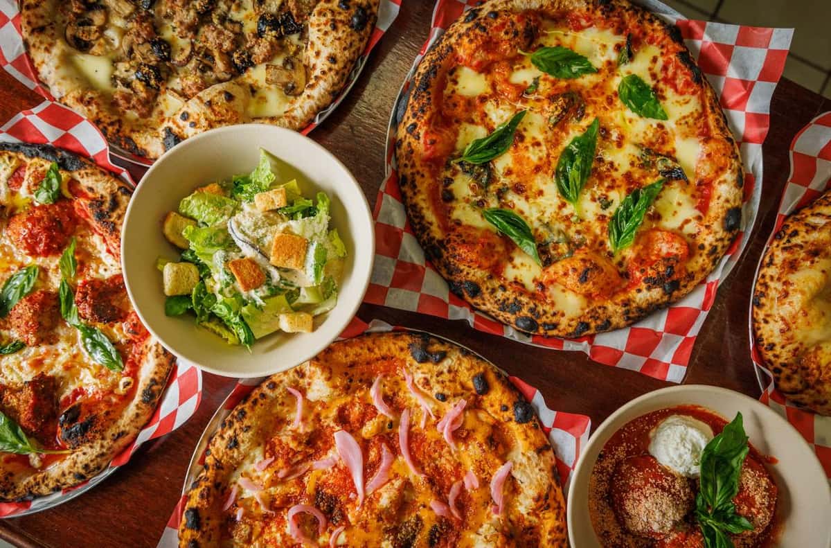 Assorted Neapolitan pizzas with fresh basil, Caesar salad, and meatballs on a rustic table with checkered liners