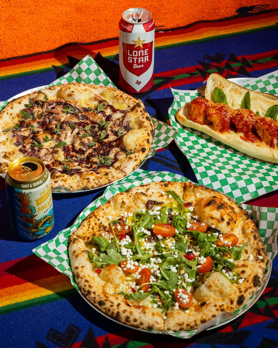 Wood-fired artisan pizzas topped with arugula, cherry tomatoes, and cheese alongside a meat-topped pizza and saucy meatball sub, served with craft beer cans on a colorful patterned tablecloth.