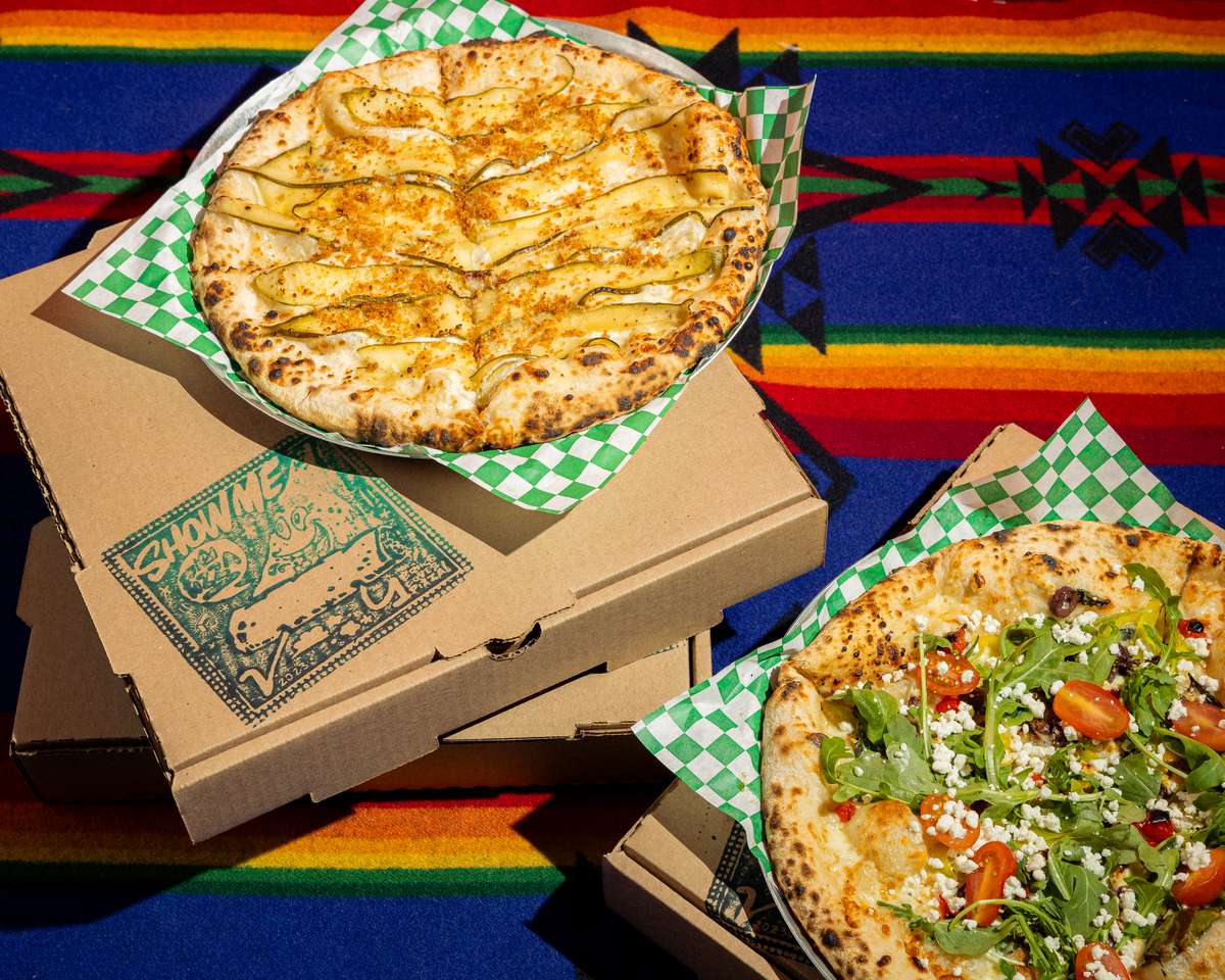 Artisan wood-fired pizzas with fresh toppings—one topped with thinly sliced vegetables and cheese, the other with arugula, cherry tomatoes, and crumbled cheese—served in takeout boxes on a colorful patterned tablecloth.