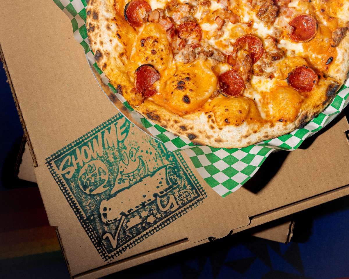 Wood-fired pizza topped with pepperoni, sausage, and melted cheese on a blistered crust over colorful textile backdrop