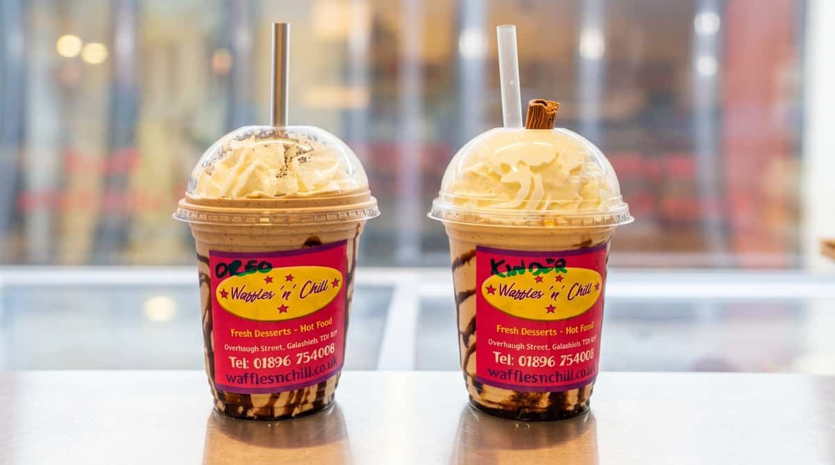 Two milkshakes with whipped cream, chocolate drizzle, and branding labels, one topped with a chocolate bar.