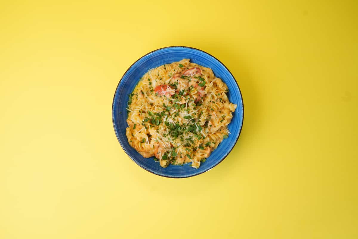 Top view of creamy fusilli pasta with tomatoes and parsley in a blue bowl, placed on a vibrant yellow background.