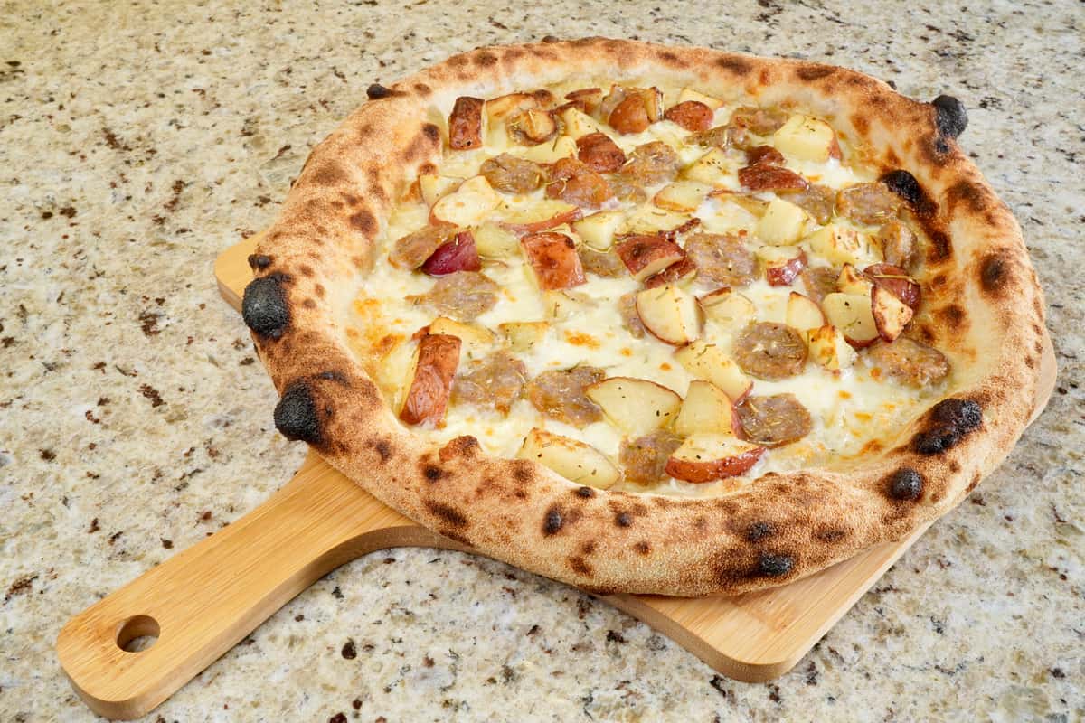 Close-up of wood-fired pizza topped with sausage, roasted potatoes, and mozzarella on a granite countertop