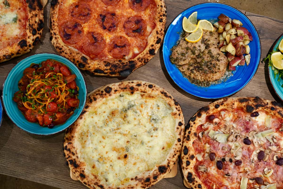 Top view of assorted Italian pizzas including pepperoni, four cheese, and prosciutto with mushrooms, accompanied by pasta, chicken piccata, and roasted potatoes