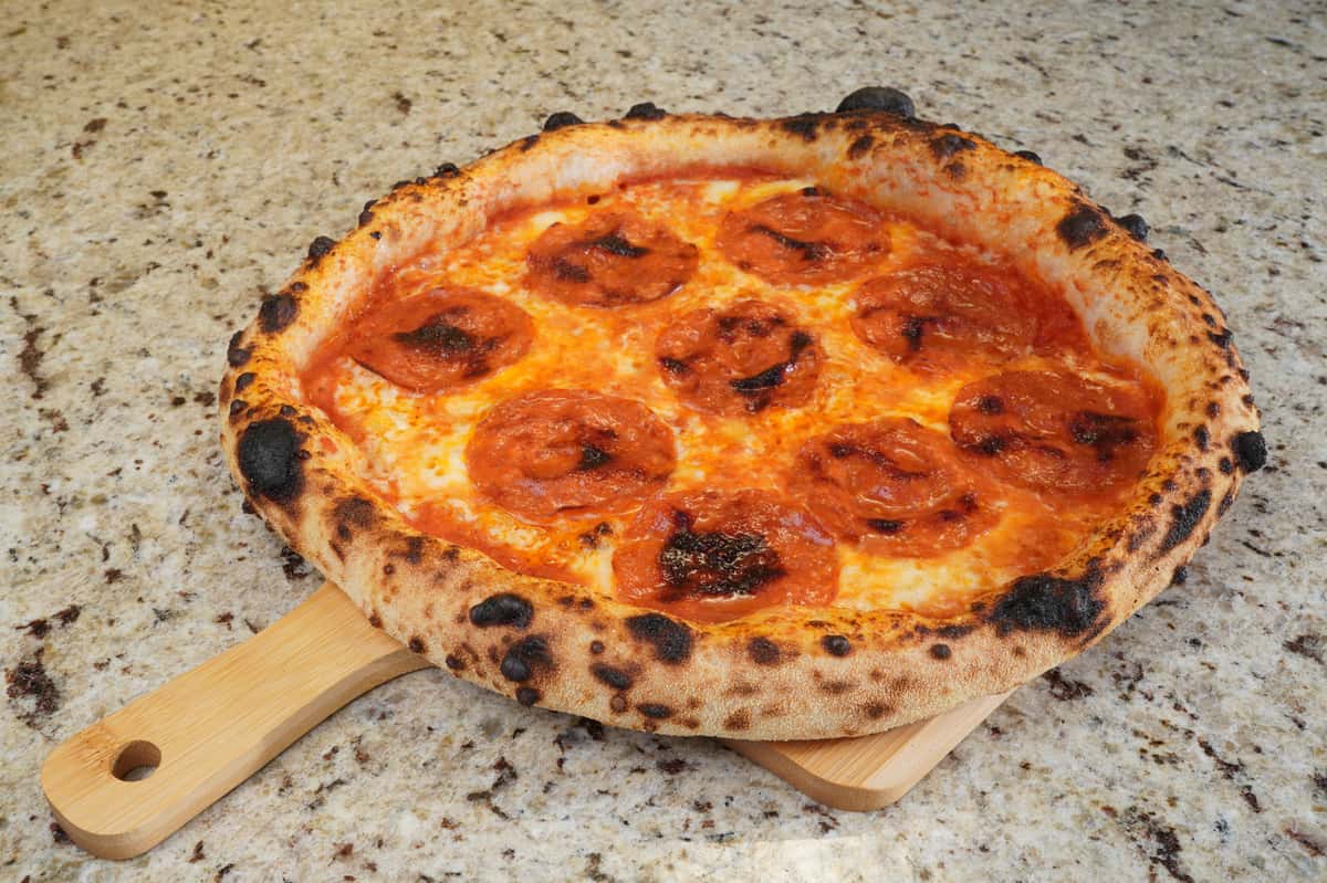 "Neapolitan-style pepperoni pizza with charred crust served on a wooden paddle over a granite countertop