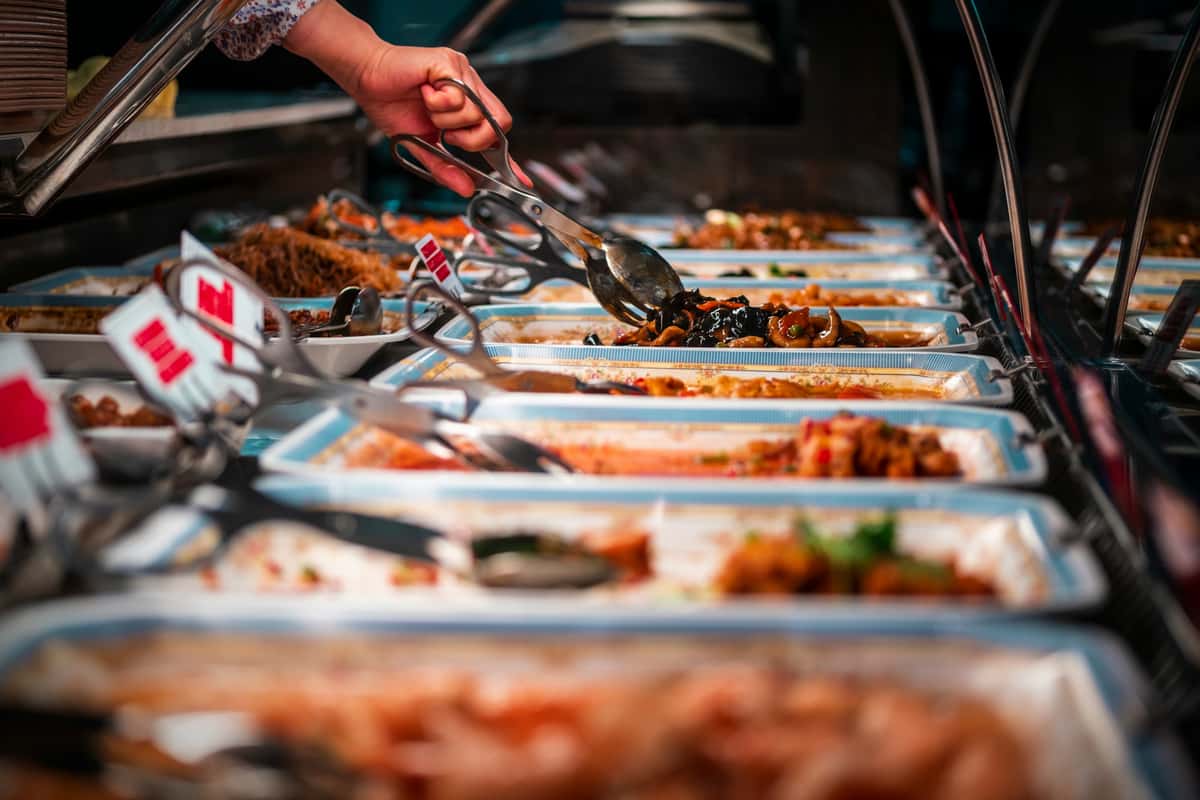Close-up of a hand serving food with tongs from a buffet line with multiple hot dishes.