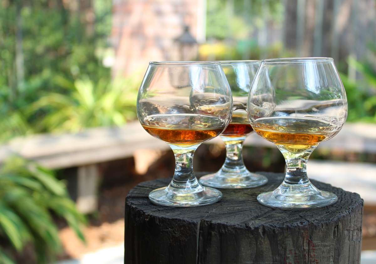 Three glass brandy snifters filled with amber-colored liquid, placed on a wooden surface with a blurred natural background.