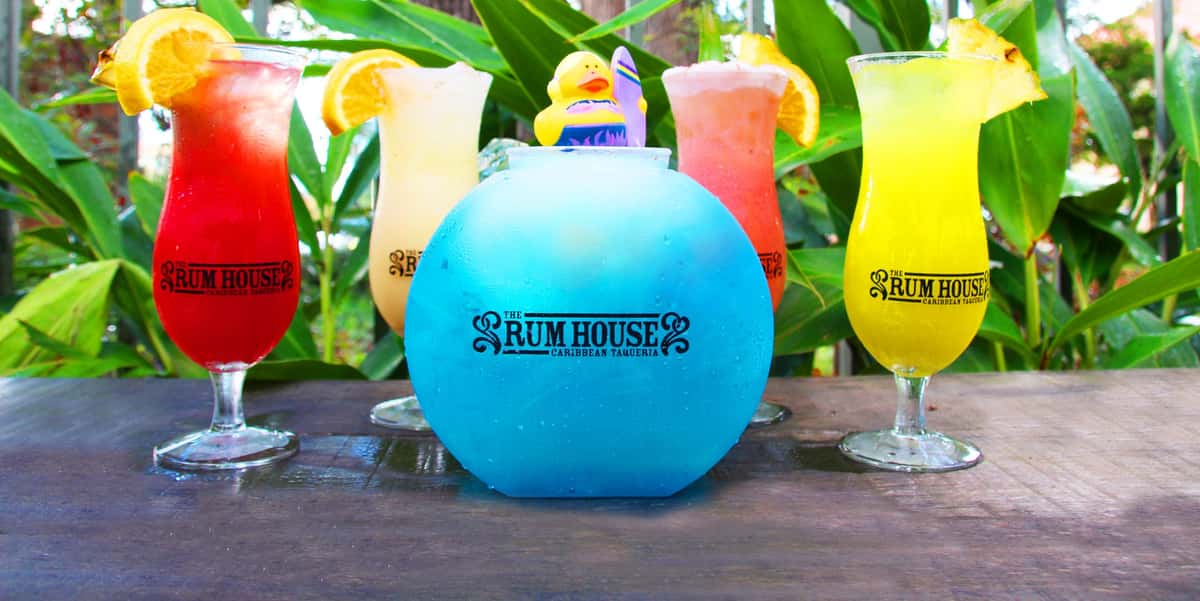 A vibrant blue drink in a large glass bowl with a rubber duck, surrounded by colorful cocktails with orange slices, all branded with "The Rum House" logo.
