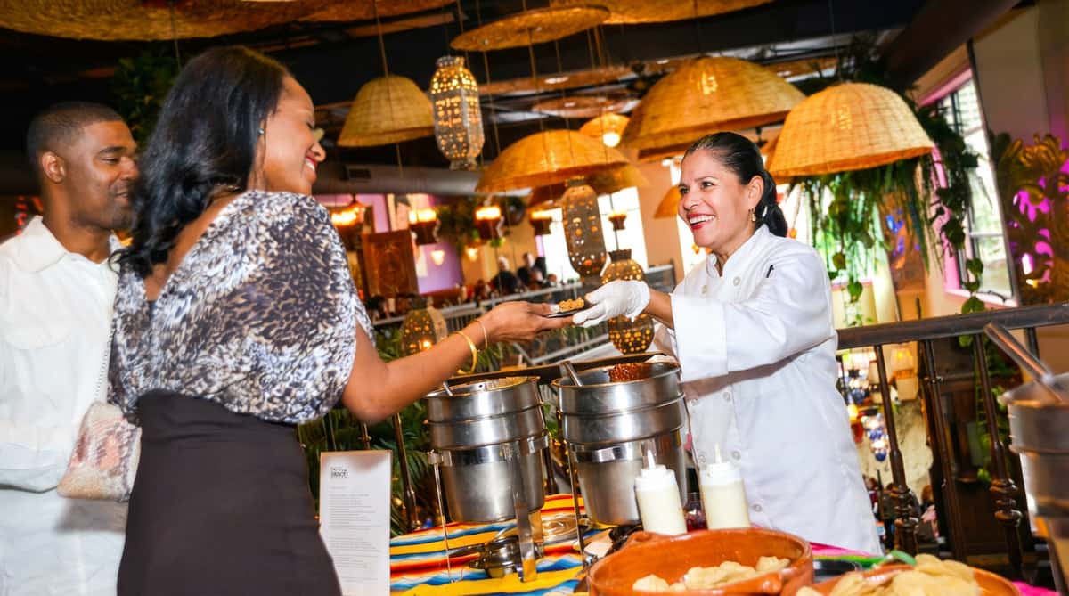 A chef in a white coat serves food on a small plate to a woman in a leopard print top from a buffet table with chip bowls and chafing dishes.