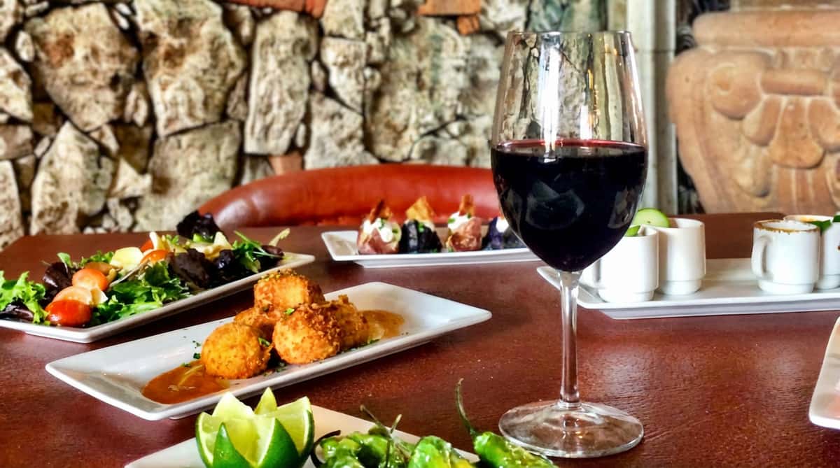 A table with a glass of red wine, plates of salad, fried appetizers, green peppers with lime, and other small dishes against a stone wall.