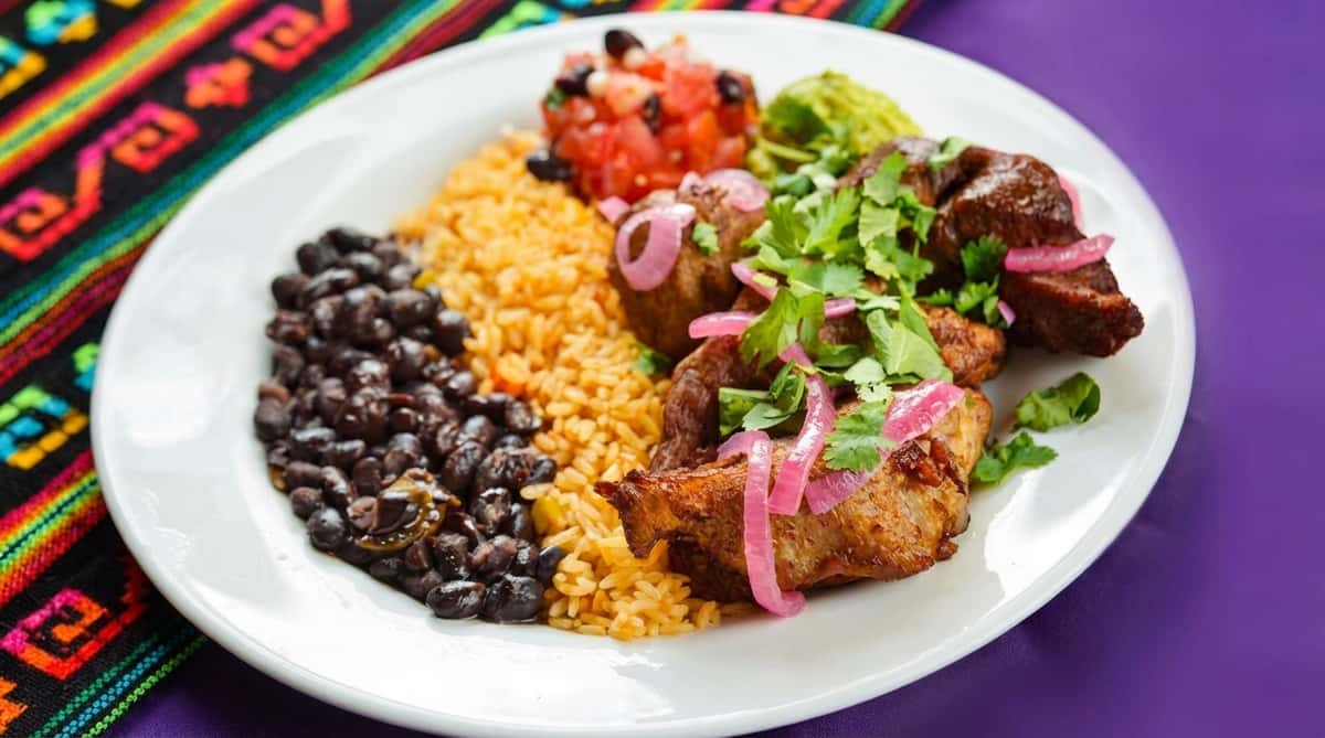 Plate with Cuban roast pork, yellow rice, black beans, pico de gallo, guacamole, and pickled red onions.