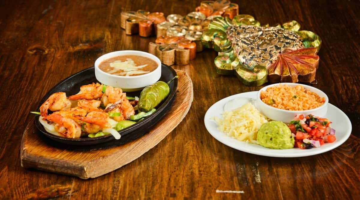 Shrimp fajitas on a sizzling platter with beans, rice, shredded cheese, guacamole, and pico de gallo.