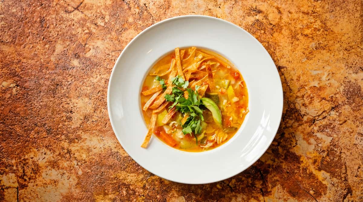 Tortilla soup with tortilla strips, cilantro, and avocado slices in a white bowl with red spice sprinkled on the rim.