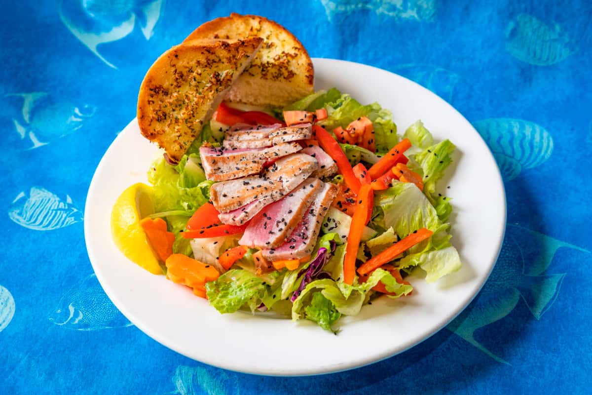 Fresh mixed greens topped with slices of sesame-crusted ahi tuna, shredded carrots, and bell peppers, served with garlic bread and a lemon wedge on a white plate against a blue nautical-patterned backdrop.