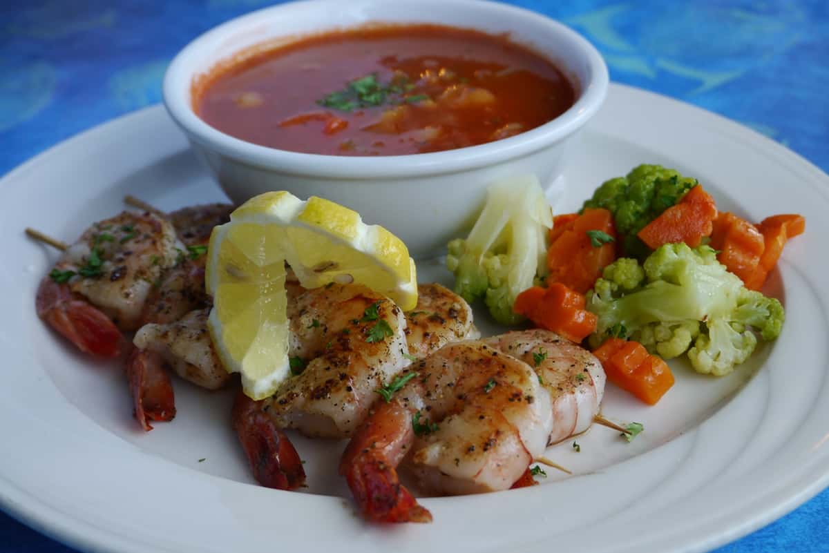 Juicy grilled shrimp skewers garnished with lemon slices, served with vegetable medley and a bowl of tomato-based seafood soup on a white plate against a blue sea-patterned tablecloth.