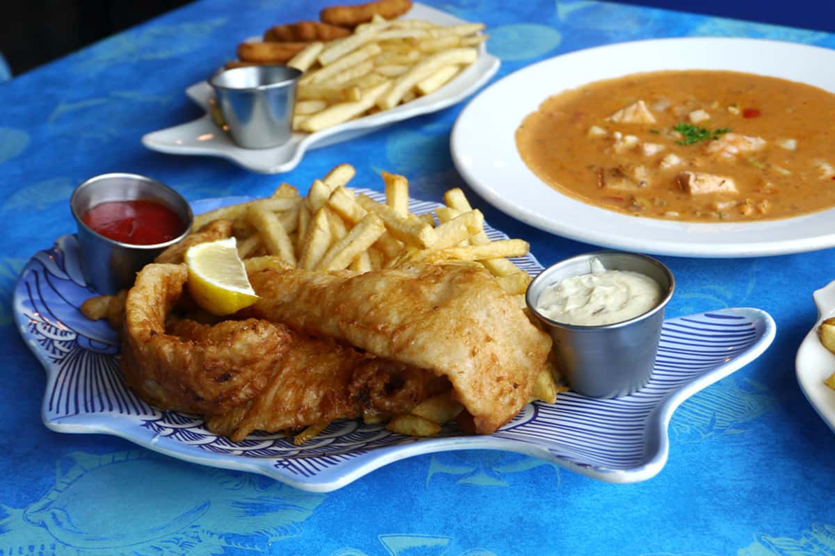 Beer-battered fish fillets with crispy fries, lemon wedge, ketchup, and tartar sauce on a fish-shaped plate, with chowder and another seafood dish in the background over a blue ocean-inspired tablecloth.